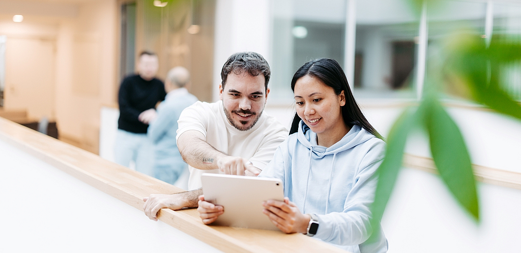 Two people are standing in an open, bright office building. They are looking at a tablet together and smiling.