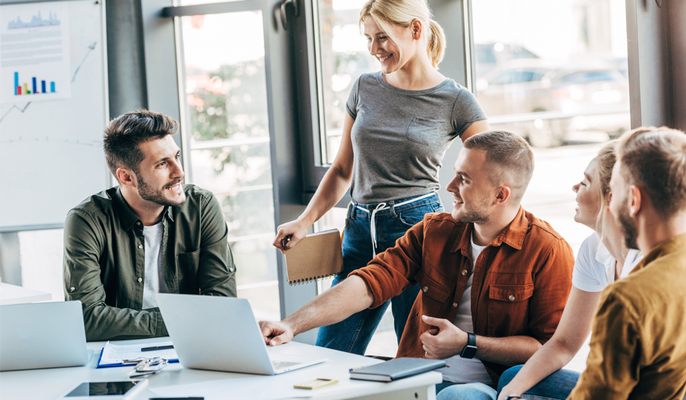 Junge Geschäftsleute in legerer Kleidung diskutieren lebhaft in einem modernen Büro bei einem Meeting. Fünf Personen sitzen oder stehen um einen Tisch mit Laptops und Unterlagen, während sie sich gegenseitig anlächeln.