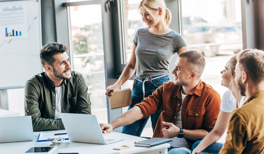 Junge Geschäftsleute in legerer Kleidung diskutieren lebhaft in einem modernen Büro bei einem Meeting. Fünf Personen sitzen oder stehen um einen Tisch mit Laptops und Unterlagen, während sie sich gegenseitig anlächeln.