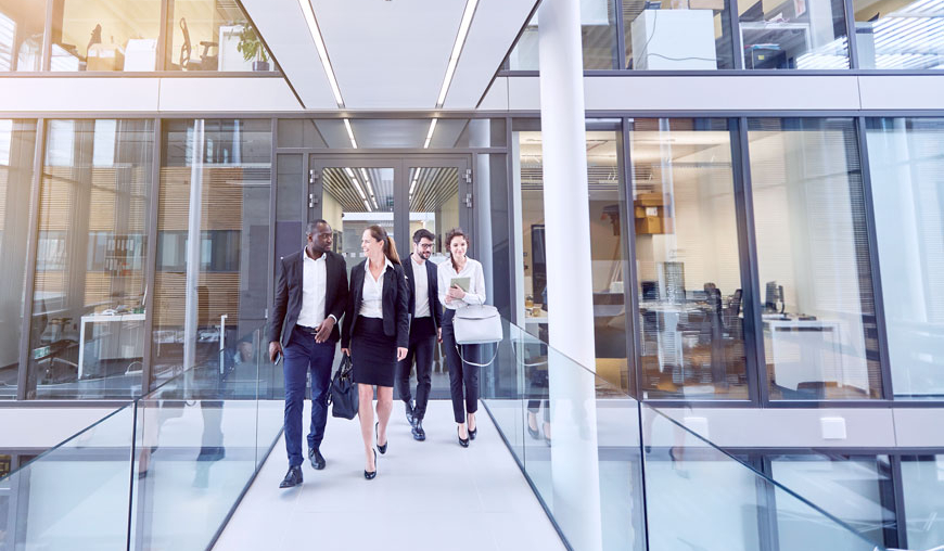 Fünf Berufstätige in Businesskleidung gehen lächelnd durch einen modernen Büroflur mit Glaswänden. Zwei Männer und drei Frauen unterhalten sich, während sie durch das lichtdurchflutete Gebäude gehen.