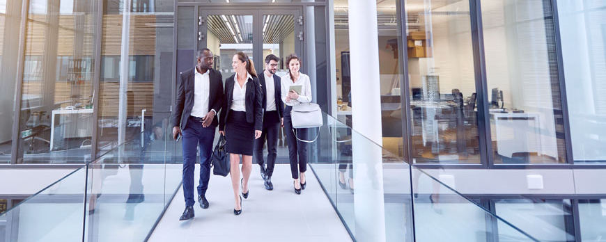 Fünf Berufstätige in Businesskleidung gehen lächelnd durch einen modernen Büroflur mit Glaswänden. Zwei Männer und drei Frauen unterhalten sich, während sie durch das lichtdurchflutete Gebäude gehen.