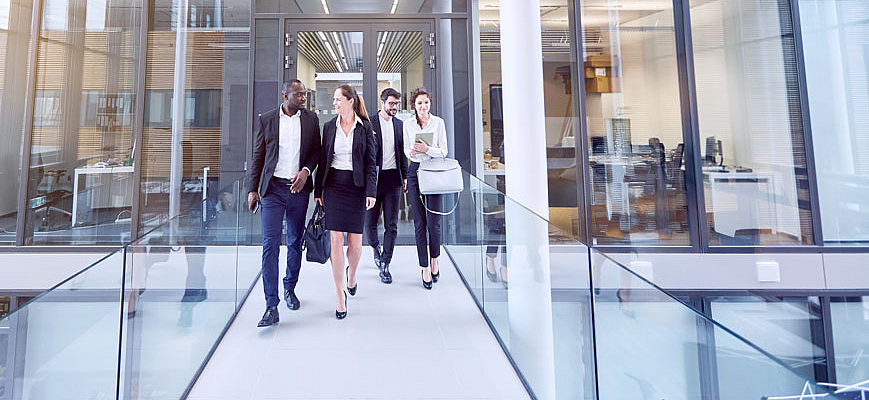 Fünf Berufstätige in Businesskleidung gehen lächelnd durch einen modernen Büroflur mit Glaswänden. Zwei Männer und drei Frauen unterhalten sich, während sie durch das lichtdurchflutete Gebäude gehen.