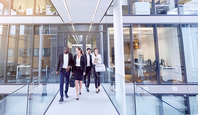 Fünf Berufstätige in Businesskleidung gehen lächelnd durch einen modernen Büroflur mit Glaswänden. Zwei Männer und drei Frauen unterhalten sich, während sie durch das lichtdurchflutete Gebäude gehen.