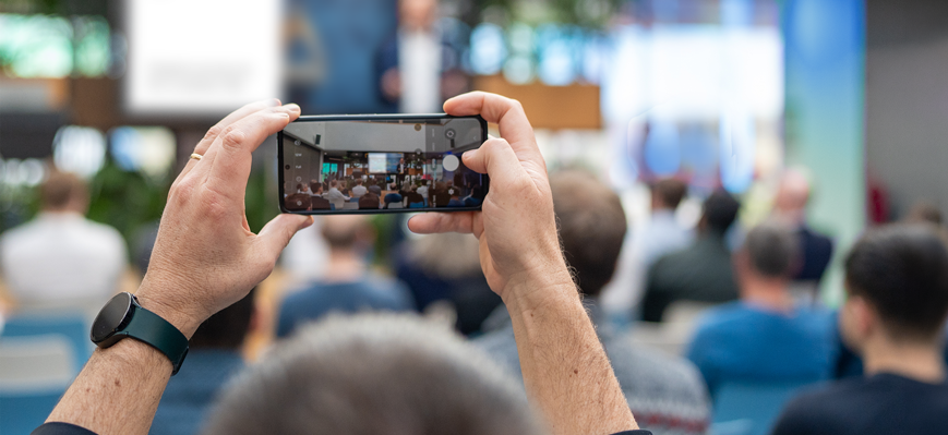 Eine Person fotografiert mit ihrem Smartphone einen Vortrag in einem Konferenzraum; im Hintergrund sitzt ein Publikum und vorne steht ein unscharfer Redner auf der Bühne.