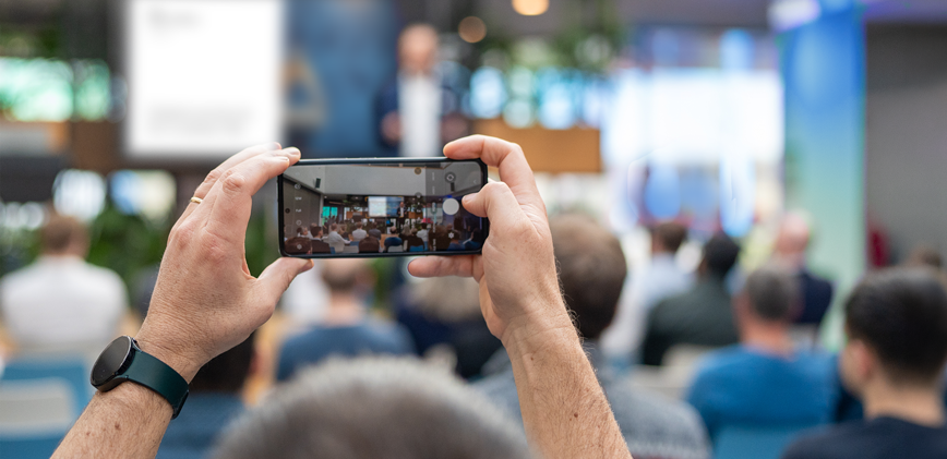 Eine Person fotografiert mit ihrem Smartphone einen Vortrag in einem Konferenzraum; im Hintergrund sitzt ein Publikum und vorne steht ein unscharfer Redner auf der Bühne.