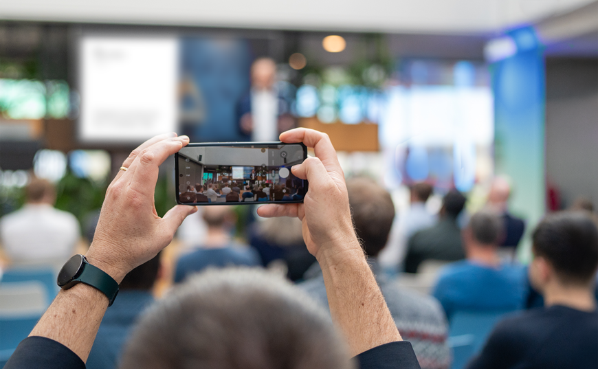 Eine Person fotografiert mit ihrem Smartphone einen Vortrag in einem Konferenzraum; im Hintergrund sitzt ein Publikum und vorne steht ein unscharfer Redner auf der Bühne.