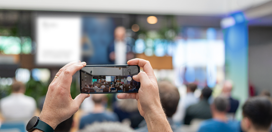 Eine Person fotografiert mit ihrem Smartphone einen Vortrag in einem Konferenzraum; im Hintergrund sitzt ein Publikum und vorne steht ein unscharfer Redner auf der Bühne.