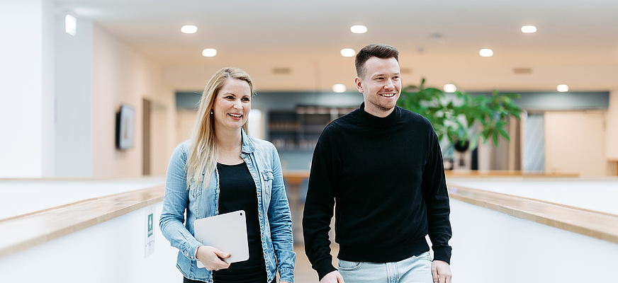 Zwei Mitarbeitende von doubleSlash gehen lächelnd durch das moderne Firmengebäude in Friedrichshafen; die Frau hält ein Tablet, beide wirken professionell, offen und im Austausch – symbolisch für Teamarbeit, Innovation und eine moderne Arbeitskultur.