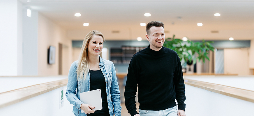 Zwei Mitarbeitende von doubleSlash gehen lächelnd durch das moderne Firmengebäude in Friedrichshafen; die Frau hält ein Tablet, beide wirken professionell, offen und im Austausch – symbolisch für Teamarbeit, Innovation und eine moderne Arbeitskultur.
