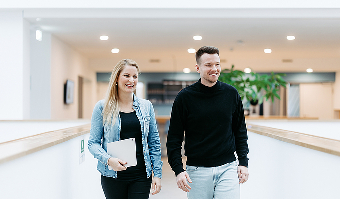 Zwei Mitarbeitende von doubleSlash gehen lächelnd durch das moderne Firmengebäude in Friedrichshafen; die Frau hält ein Tablet, beide wirken professionell, offen und im Austausch – symbolisch für Teamarbeit, Innovation und eine moderne Arbeitskultur.