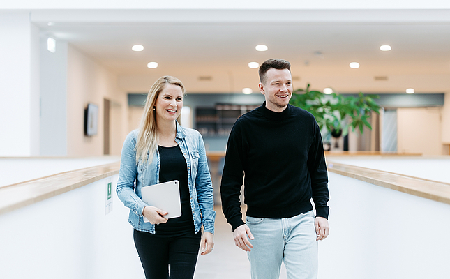 Zwei Mitarbeitende von doubleSlash gehen lächelnd durch das moderne Firmengebäude in Friedrichshafen; die Frau hält ein Tablet, beide wirken professionell, offen und im Austausch – symbolisch für Teamarbeit, Innovation und eine moderne Arbeitskultur.