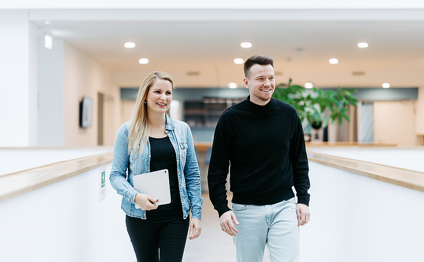 Zwei Mitarbeitende von doubleSlash gehen lächelnd durch das moderne Firmengebäude in Friedrichshafen; die Frau hält ein Tablet, beide wirken professionell, offen und im Austausch – symbolisch für Teamarbeit, Innovation und eine moderne Arbeitskultur.