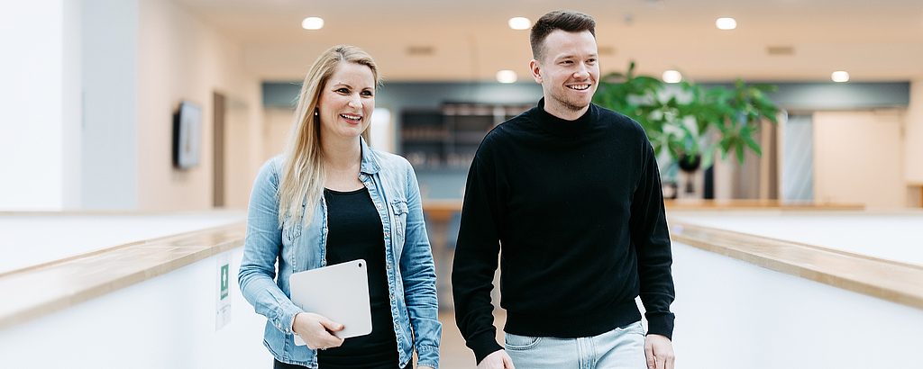 Zwei Mitarbeitende von doubleSlash gehen lächelnd durch das moderne Firmengebäude in Friedrichshafen; die Frau hält ein Tablet, beide wirken professionell, offen und im Austausch – symbolisch für Teamarbeit, Innovation und eine moderne Arbeitskultur.