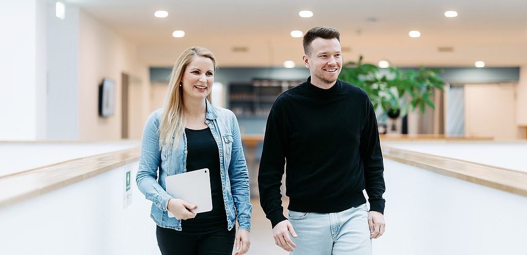 Zwei Mitarbeitende von doubleSlash gehen lächelnd durch das moderne Firmengebäude in Friedrichshafen; die Frau hält ein Tablet, beide wirken professionell, offen und im Austausch – symbolisch für Teamarbeit, Innovation und eine moderne Arbeitskultur.