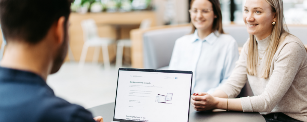 Drei Personen sitzen an einem Tisch und besprechen sich. Wir schauen einem Mann über die Schulter. Er hat einen Laptop vor sich und nutzt Secure Password Share.