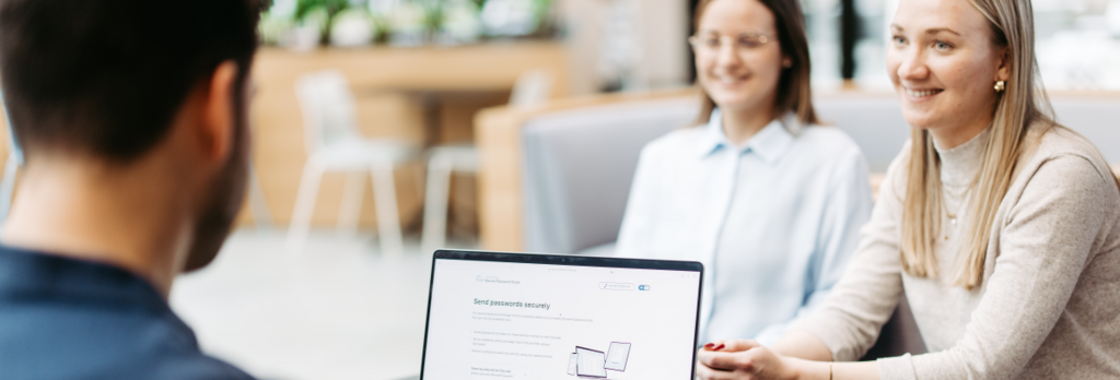 Drei Personen sitzen an einem Tisch und besprechen sich. Wir schauen einem Mann über die Schulter. Er hat einen Laptop vor sich und nutzt Secure Password Share.