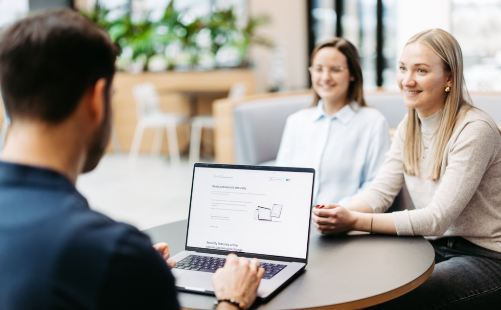 Three people are sitting at a table discussing something. We look over a man's shoulder. He has a laptop in front of him and is using Secure Password Share.