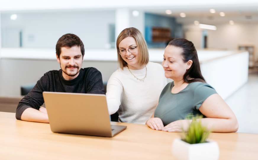 Three employees are standing in the coffee kitchen, looking at a laptop together and discussing things.