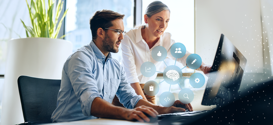 Two people are working together at a computer, with a graphic above them showing a connected brain symbol and icons for data, analytics, and digital processes.