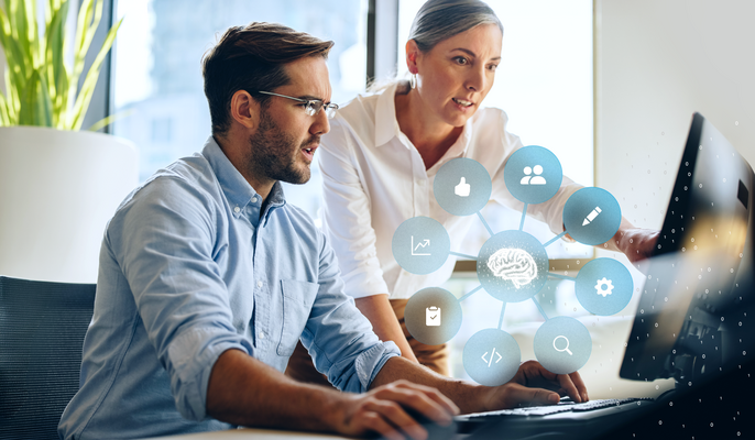 Two people are working together at a computer, with a graphic above them showing a connected brain symbol and icons for data, analytics, and digital processes.
