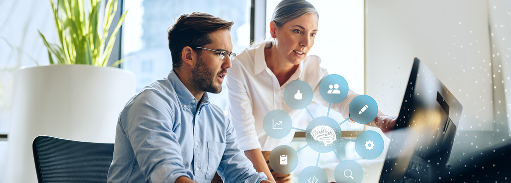 Two people are working together at a computer, with a graphic above them showing a connected brain symbol and icons for data, analytics, and digital processes.