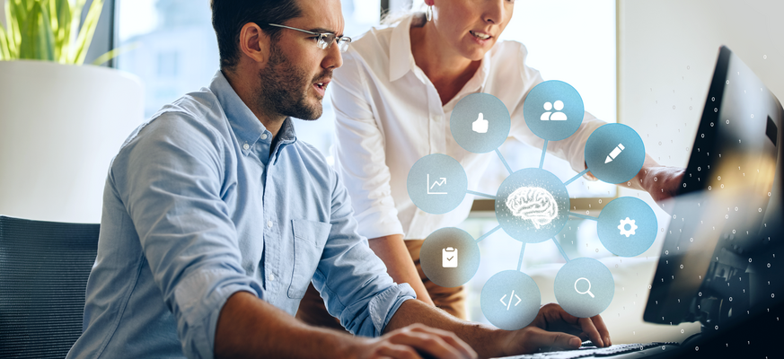 Zwei Personen arbeiten gemeinsam am Computer; darüber eine Grafik mit vernetztem Gehirn-Symbol und Icons für Daten, Analyse und digitale Prozesse.