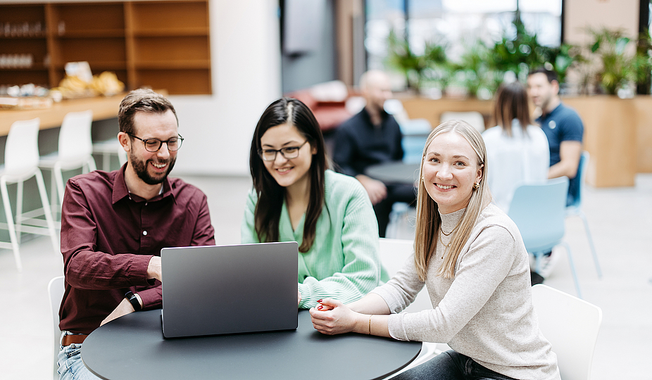 Drei Menschen schauen gemeinsam auf einen Laptop, eine Frau schaut direkt in die Kamera. Sie befinden sich in einem großen, hellen, freundlichen Atrium. Im Hintergrund besprechen sich noch weitere Menschen.
