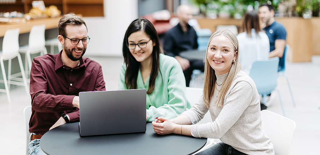 Drei Menschen schauen gemeinsam auf einen Laptop, eine Frau schaut direkt in die Kamera. Sie befinden sich in einem großen, hellen, freundlichen Atrium. Im Hintergrund besprechen sich noch weitere Menschen.