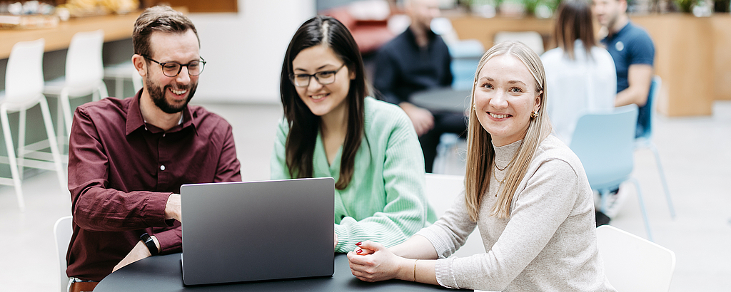 Three people are looking at a laptop together, one woman is looking directly into the camera. They are in a large, bright, friendly atrium. Other people are talking in the background.
