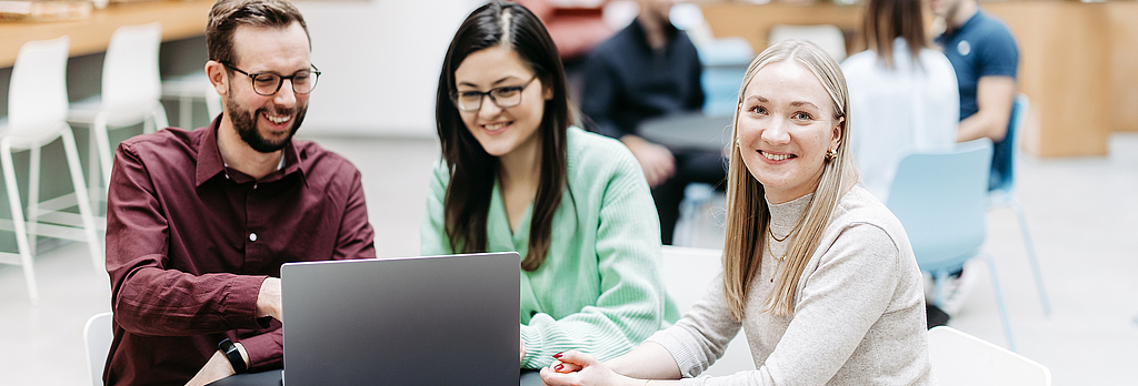 Three people are looking at a laptop together, one woman is looking directly into the camera. They are in a large, bright, friendly atrium. Other people are talking in the background.