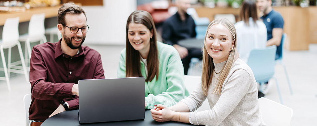 Three people are looking at a laptop together, one woman is looking directly into the camera. They are in a large, bright, friendly atrium. Other people are talking in the background.