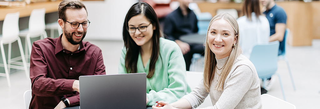 Three people are looking at a laptop together, one woman is looking directly into the camera. They are in a large, bright, friendly atrium. Other people are talking in the background.