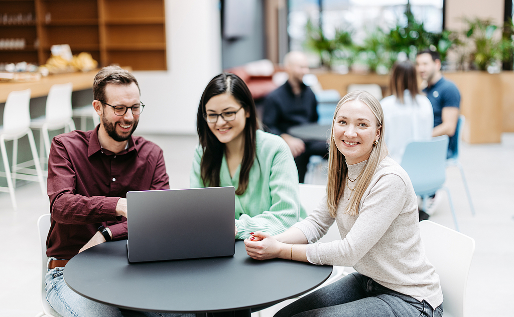 Drei Menschen schauen gemeinsam auf einen Laptop, eine Frau schaut direkt in die Kamera. Sie befinden sich in einem großen, hellen, freundlichen Atrium. Im Hintergrund besprechen sich noch weitere Menschen.