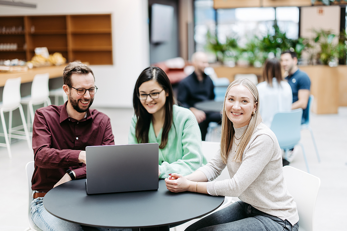 Drei Menschen schauen gemeinsam auf einen Laptop, eine Frau schaut direkt in die Kamera. Sie befinden sich in einem großen, hellen, freundlichen Atrium. Im Hintergrund besprechen sich noch weitere Menschen.