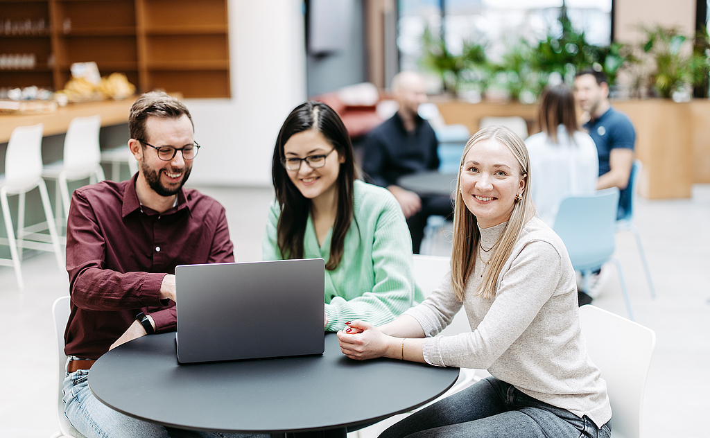 Drei Menschen schauen gemeinsam auf einen Laptop, eine Frau schaut direkt in die Kamera. Sie befinden sich in einem großen, hellen, freundlichen Atrium. Im Hintergrund besprechen sich noch weitere Menschen.