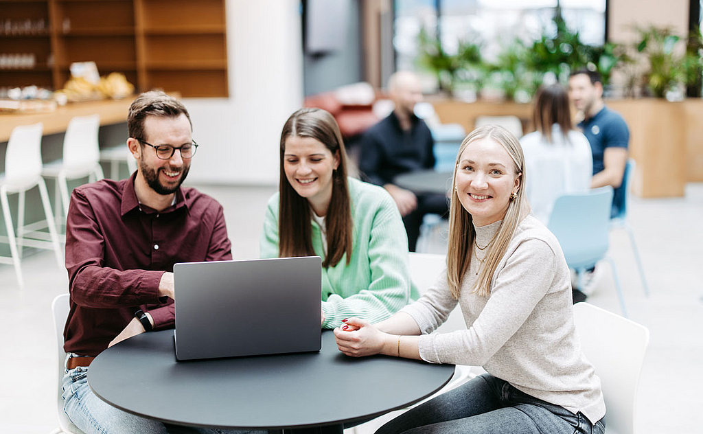 Drei Menschen schauen gemeinsam auf einen Laptop, eine Frau schaut direkt in die Kamera. Sie befinden sich in einem großen, hellen, freundlichen Atrium. Im Hintergrund besprechen sich noch weitere Menschen.