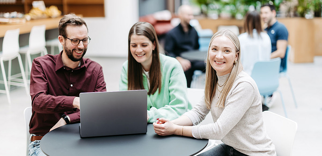 Drei Menschen schauen gemeinsam auf einen Laptop, eine Frau schaut direkt in die Kamera. Sie befinden sich in einem großen, hellen, freundlichen Atrium. Im Hintergrund besprechen sich noch weitere Menschen.