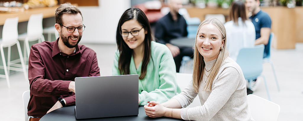 Drei Menschen schauen gemeinsam auf einen Laptop, eine Frau schaut direkt in die Kamera. Sie befinden sich in einem großen, hellen, freundlichen Atrium. Im Hintergrund besprechen sich noch weitere Menschen.