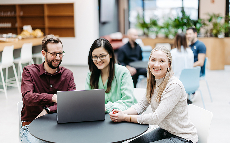 Drei Menschen schauen gemeinsam auf einen Laptop, eine Frau schaut direkt in die Kamera. Sie befinden sich in einem großen, hellen, freundlichen Atrium. Im Hintergrund besprechen sich noch weitere Menschen.
