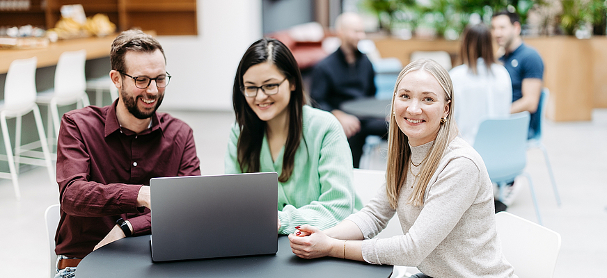 Drei Menschen schauen gemeinsam auf einen Laptop, eine Frau schaut direkt in die Kamera. Sie befinden sich in einem großen, hellen, freundlichen Atrium. Im Hintergrund besprechen sich noch weitere Menschen. – KI-generiertes Bild