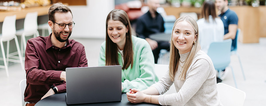 Drei Menschen schauen gemeinsam auf einen Laptop, eine Frau schaut direkt in die Kamera. Sie befinden sich in einem großen, hellen, freundlichen Atrium. Im Hintergrund besprechen sich noch weitere Menschen.