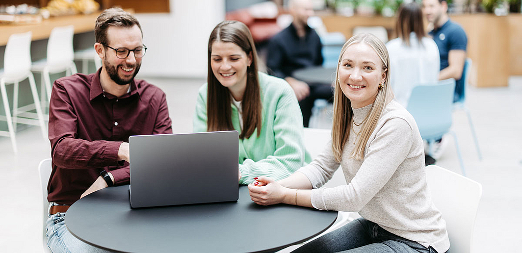 Drei Menschen schauen gemeinsam auf einen Laptop, eine Frau schaut direkt in die Kamera. Sie befinden sich in einem großen, hellen, freundlichen Atrium. Im Hintergrund besprechen sich noch weitere Menschen.