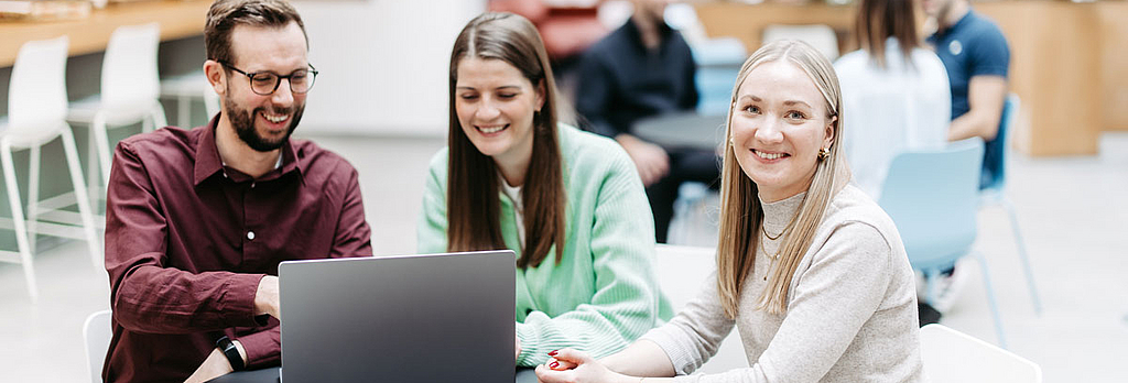 Drei Menschen schauen gemeinsam auf einen Laptop, eine Frau schaut direkt in die Kamera. Sie befinden sich in einem großen, hellen, freundlichen Atrium. Im Hintergrund besprechen sich noch weitere Menschen.