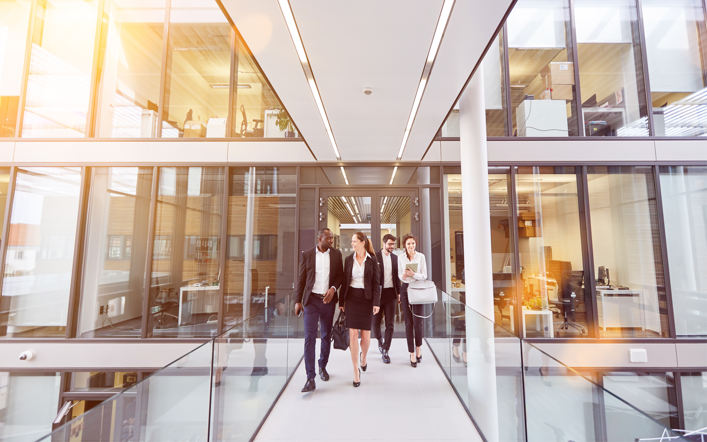 Fünf Berufstätige in Businesskleidung gehen lächelnd durch einen modernen Büroflur mit Glaswänden. Zwei Männer und drei Frauen unterhalten sich, während sie durch das lichtdurchflutete Gebäude gehen.