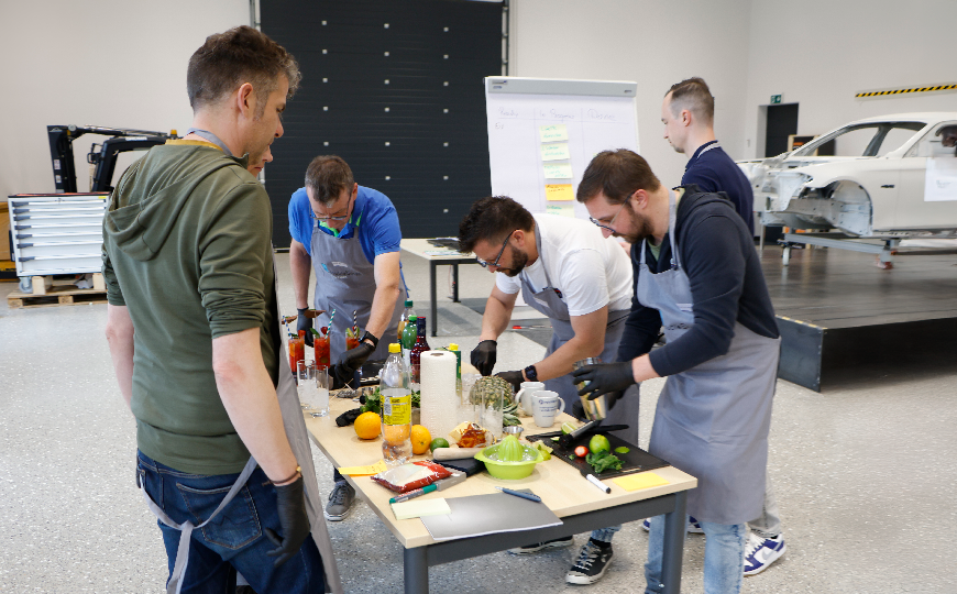 Mehrere Personen in Schürzen bereiten gemeinsam Getränke und Zutaten an einem Tisch in einer Werkhalle vor.