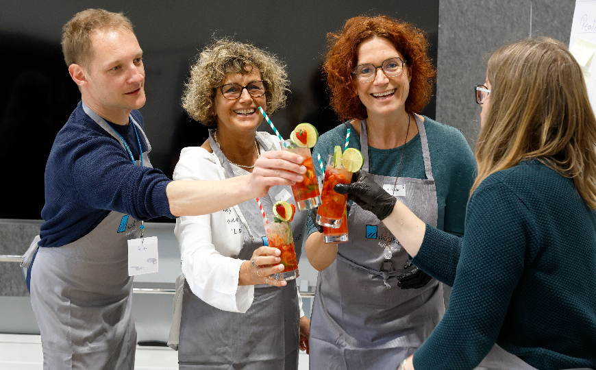 Vier Personen in Schürzen stoßen lächelnd mit bunten Cocktails an und feiern gemeinsam das Ergebnis ihrer Teamarbeit.