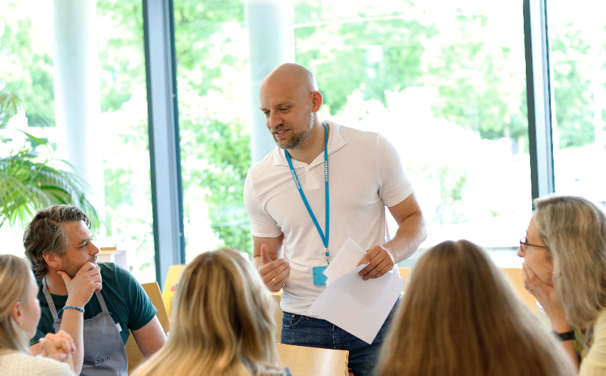 Ein Mann spricht gestikulierend zu einer sitzenden Gruppe in hellem Raum, hält Notizen in der Hand und moderiert eine gemeinsame Besprechung.