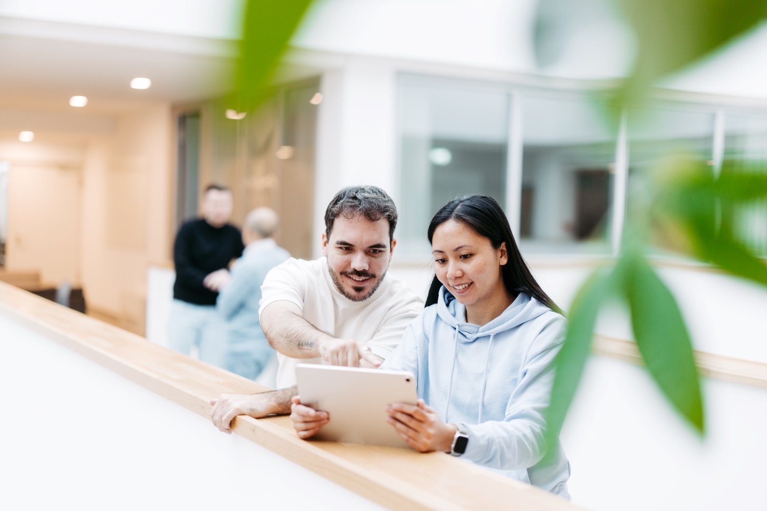 Two people are standing in an open, bright office building. They are looking at a tablet together and smiling.