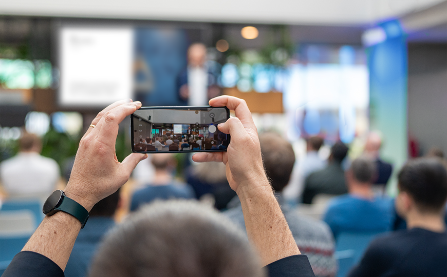 Eine Person fotografiert mit ihrem Smartphone einen Vortrag in einem Konferenzraum; im Hintergrund sitzt ein Publikum und vorne steht ein unscharfer Redner auf der Bühne.